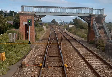 Forward-facing video on the AIVR Platform showing a rural station approach with a footbridge, switches and crossings, and a virtual signal overlaid on the imagery with a yellow measurement line indicating sighting distance.