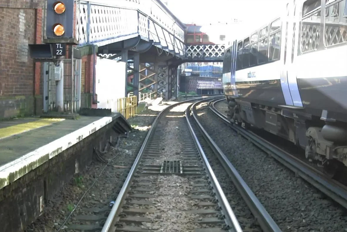 Forward-facing video from the AIVR Platform showing an urban station approach with platforms, signals, an overhead footbridge, and a train on an adjacent track, used to support station dwell time analysis and operational review.