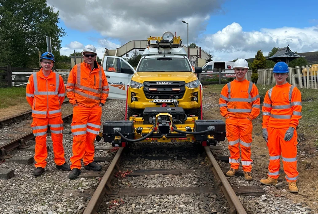 Network Rail and OBC team members in orange hi-vis PPE standing on a rail track beside a Network Rail road-rail vehicle (RRV) with an AIVR survey trailer attached.