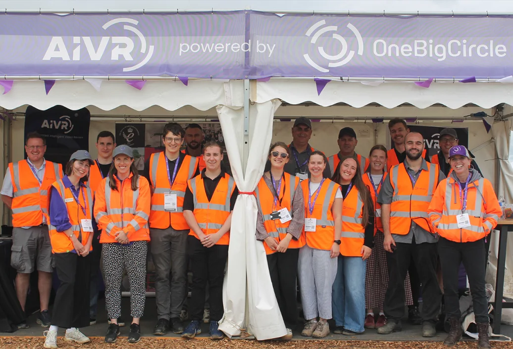 The One Big Circle team standing together under the AIVR stand awning at Rail Live 2026, wearing orange hi-vis vests and event lanyards.