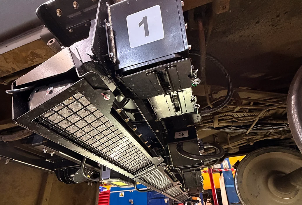Underbody view of a train showing black camera housings and a mesh-covered LED lighting array mounted above the rail.