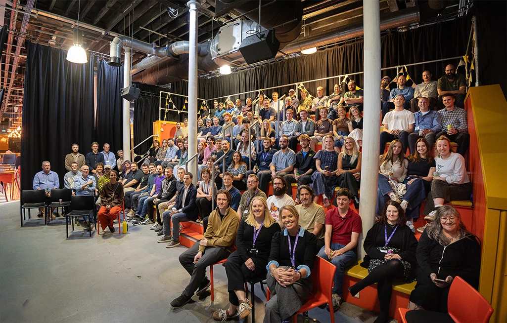 A large group of the OBC team sitting together on tiered orange and yellow seating and chairs in an industrial-style venue decorated with bunting, smiling at the camera.