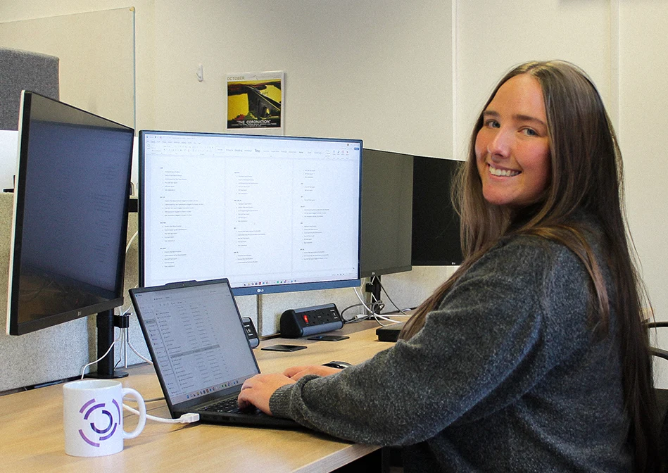 Eliza, a commercial and quality associate, smiles over her shoulder while working at a laptop and monitor in the OBC office.