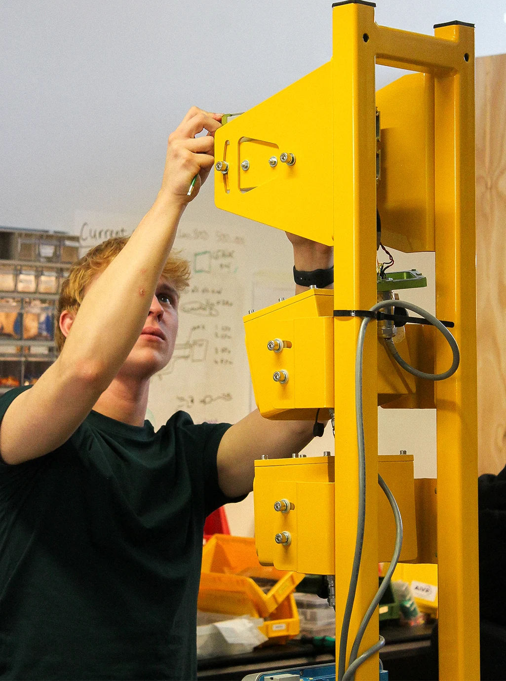 James, a hardware engineer, fixes a component onto a yellow mounting frame holding camera enclosures in the OBC lab.