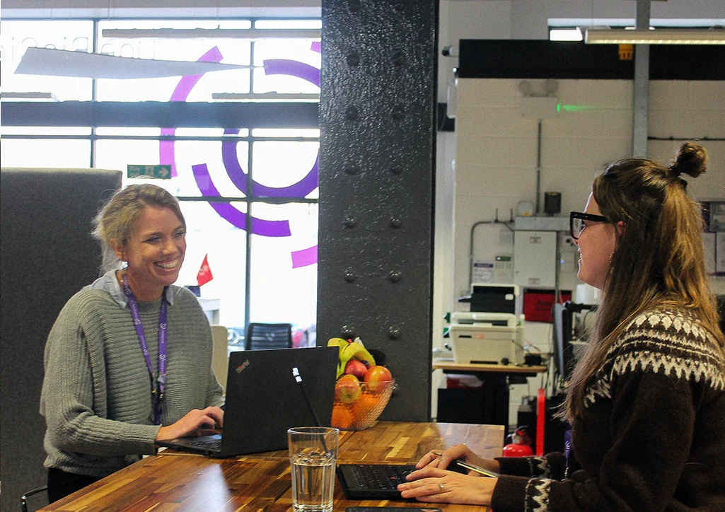 Two OBC colleagues laugh and chat while working on laptops at a shared wooden desk, with the OBC logo on the office window behind them.