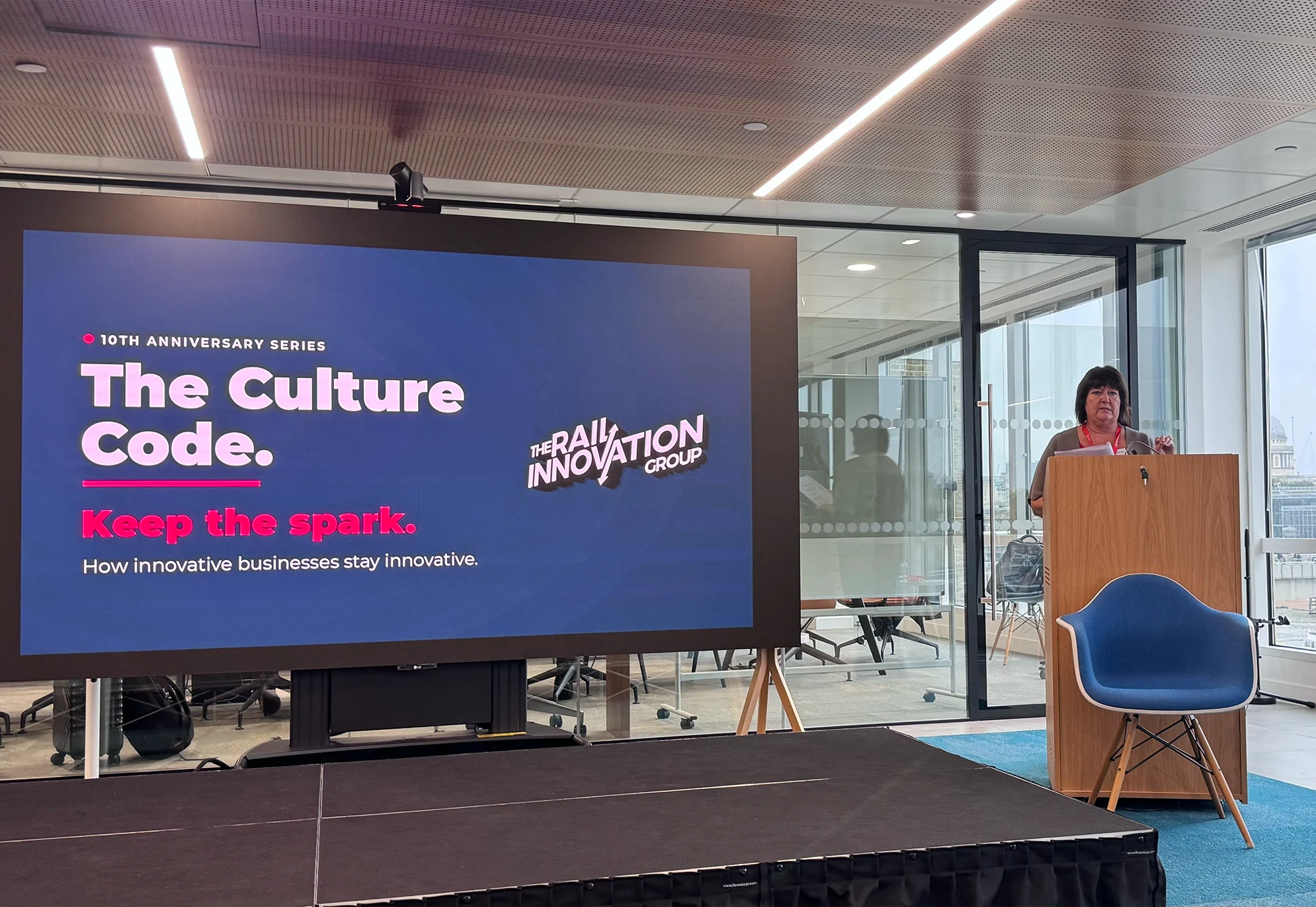 Speaker at a lectern next to a screen showing “The Culture Code. Keep the spark.” with The Rail Innovation Group logo.