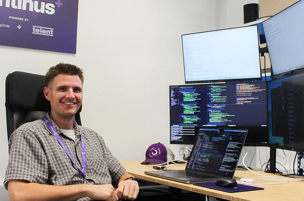 Harvey, a software engineer, smiles at the camera from his desk in the OBC office, with code on several monitors and a laptop beside him.