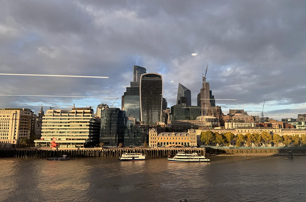 View across the River Thames to the City of London skyline under a dramatic sky, with boats moored along the river.